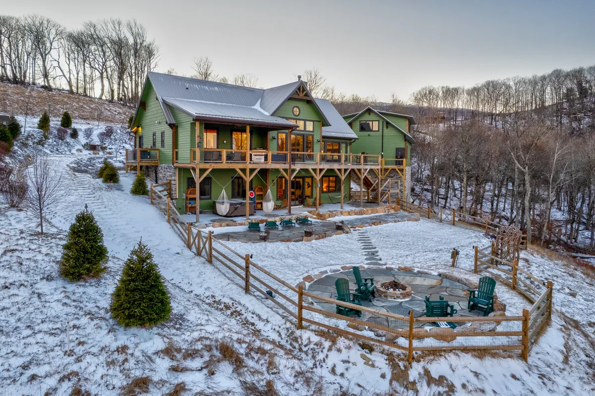 The image shows the green house with a welcoming outdoor seating area and a fire pit set against the backdrop of the tranquil Blue Ridge environment. The fire pit is surrounded by seating, inviting families to gather, and the deck offers views of the surrounding nature.