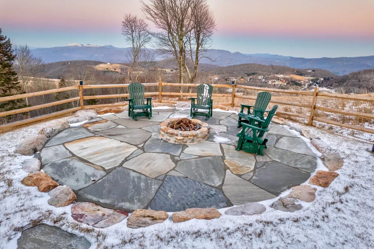 An inviting exterior scene featuring a stone fire pit surrounded by Adirondack chairs on a stone patio. The mountains in the background glow with sunset hues, and the space looks perfect for relaxing outdoors with friends, stargazing, or enjoying the views after a day of eco-friendly adventures.