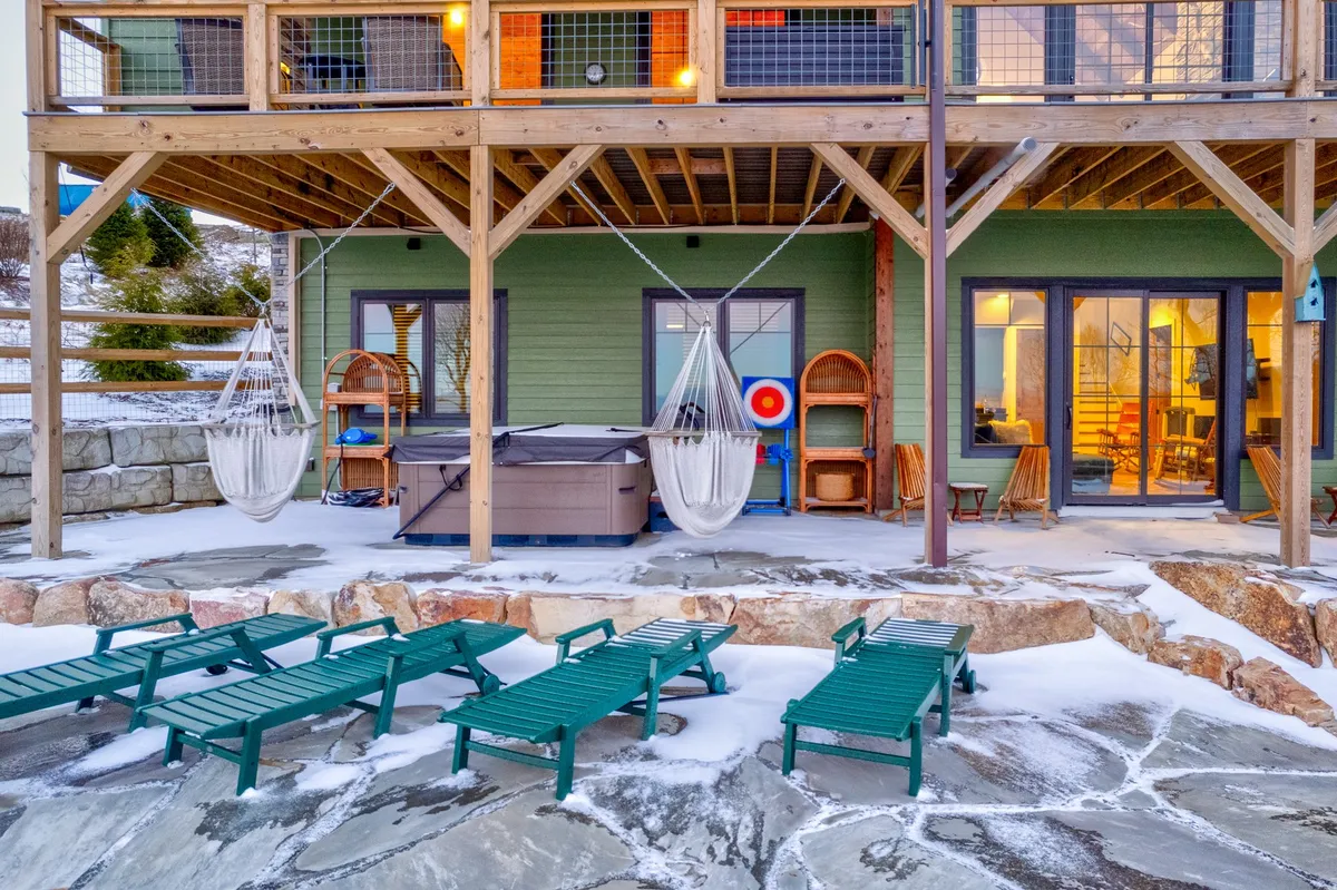 This exterior image spotlights the hot tub, hammock chairs, lounge chairs, and large windows—the perfect setting for relaxation after a day exploring the mountains. The wooden beams and natural surroundings reinforce the cabin’s mountain setting.