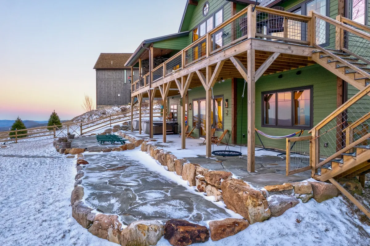A picturesque view of the green house exterior featuring a wooden deck and stone patio, all set against a snowy mountain landscape. The image suggests opportunities for wintertime outdoor relaxation and scenic appreciation just steps from the house.