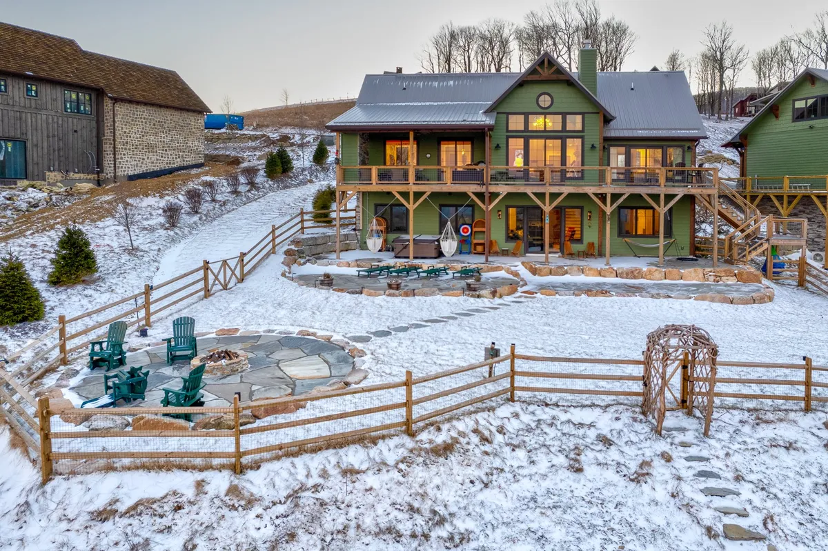 This exterior image features the green house, a stone fire pit with seating, and a snow-covered landscape. The inviting setup is perfect for relaxing outdoors, with space for pets and owners to enjoy the fresh air together. The combination of open space and gathering spots encourages outdoor activities and pet-friendly bonding.