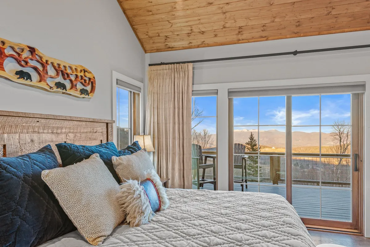 A tranquil king bedroom featuring a wooden bed frame with plush bedding, decorative wall art, and large windows that frame a breathtaking mountain view. Soft, natural light fills the room, enhancing its serene atmosphere.
