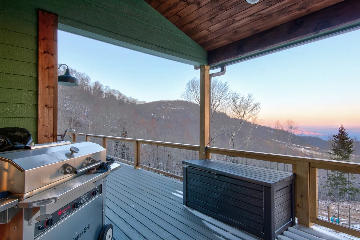 A deck area featuring a built-in grill, deck railing, and a panoramic mountain view. The scene is set for outdoor dining or a barbecue, inviting guests to savor meals in the fresh mountain air.