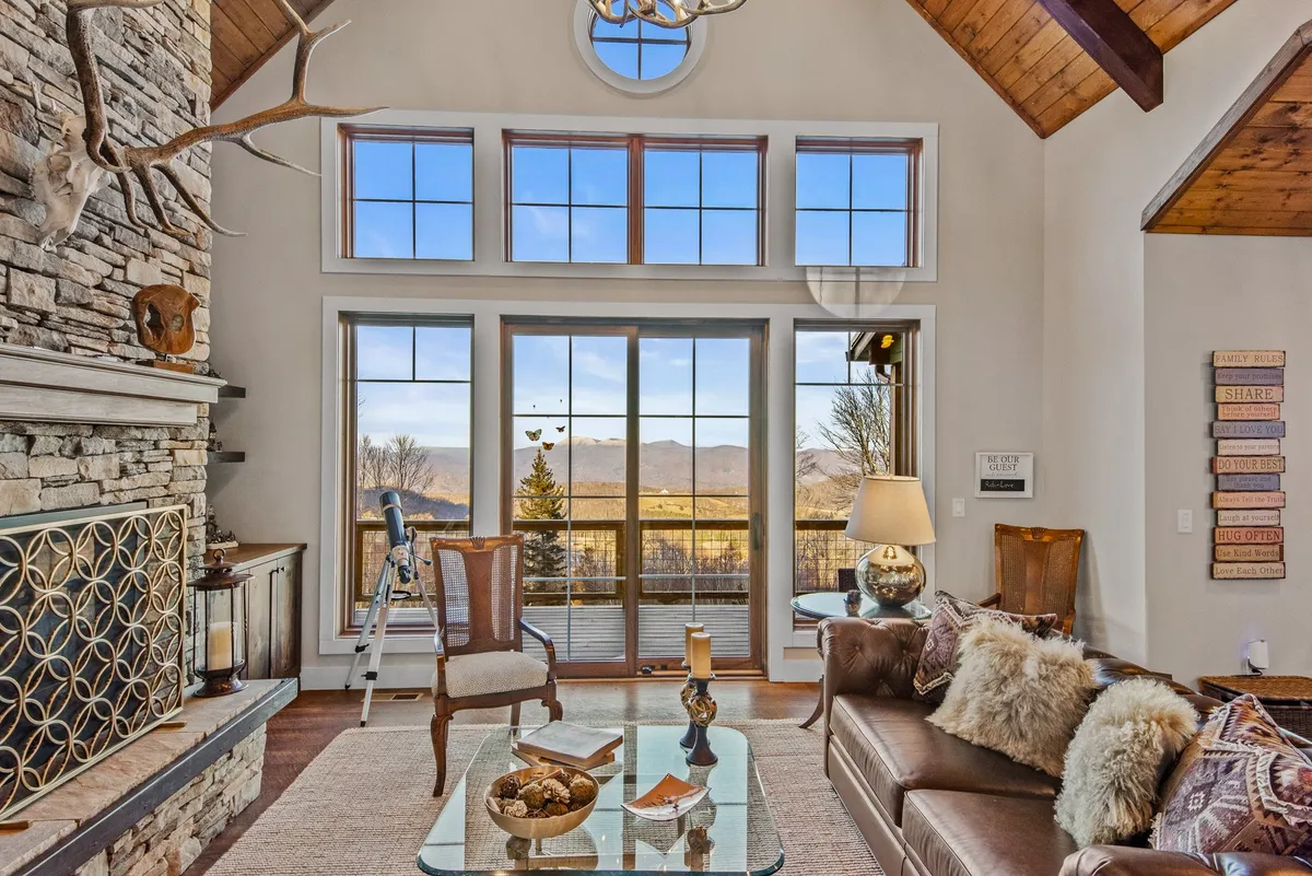 This image showcases a great room with a striking stone fireplace, high wooden beams, large windows framing the mountain views, and stylish, comfortable furniture. The natural light pours in, highlighting both the cozy seating area and the stunning scenery outside.