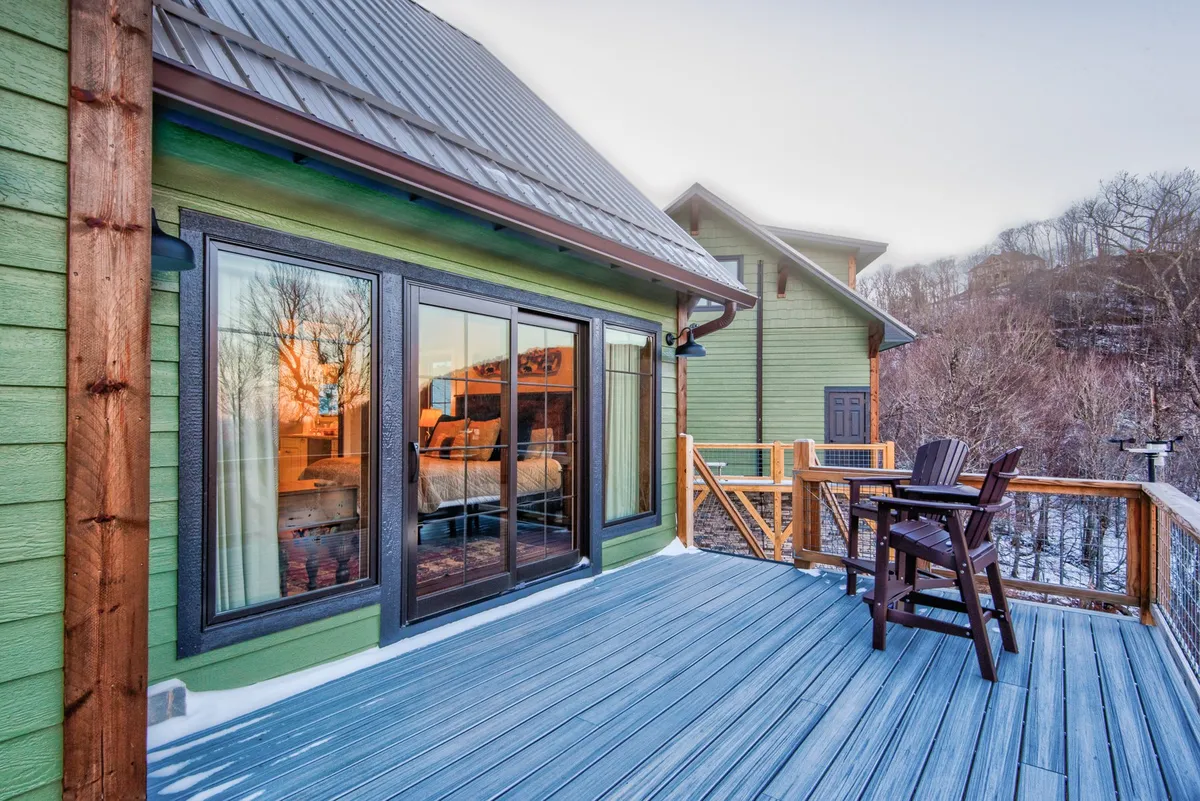 A serene outdoor seating area is showcased on a wooden deck, complete with large glass windows that reflect the surrounding scenery. The space invites guests to gather, relax with a drink, or take in the sunset, perfectly complementing the retreat vibe.