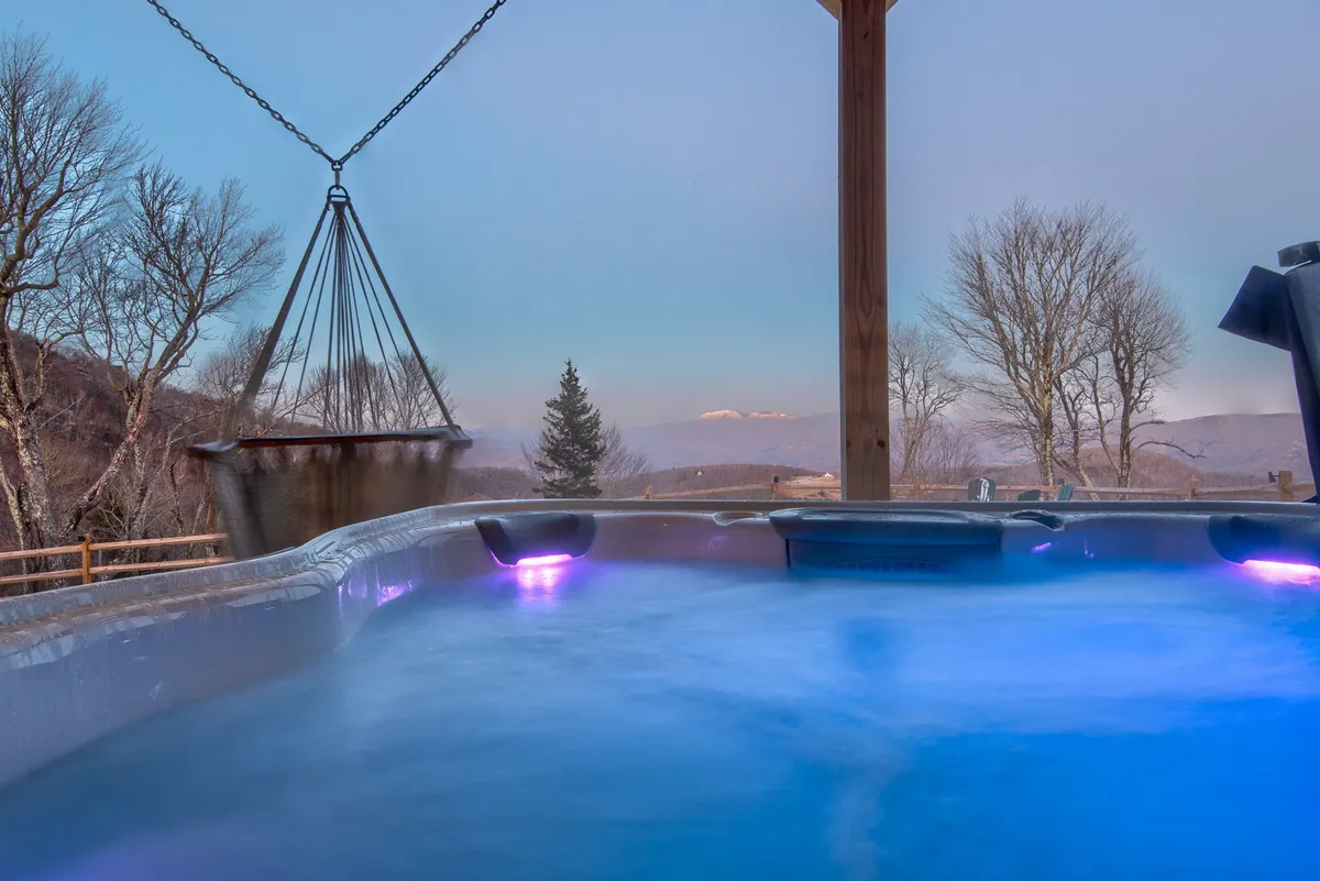 This image features a bubbling hot tub set on a patio, with a hanging chair providing a cozy spot to relax. The backdrop includes trees and sweeping mountain views, capturing a tranquil outdoor oasis.