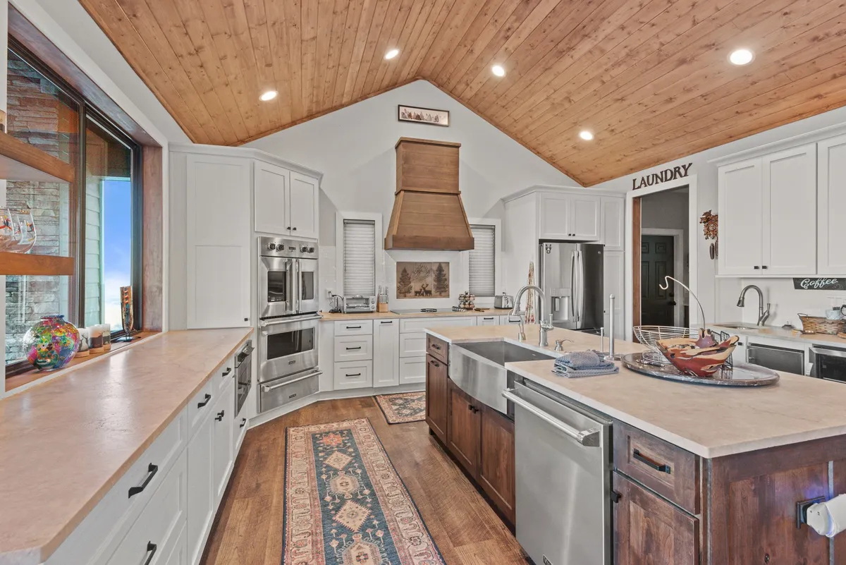 This image features a warm, welcoming kitchen with a central island counter, gleaming stainless steel appliances, wooden ceiling, and a cozy rug. The space is clearly designed for cooking and entertaining, with ample workspace and modern amenities visible.